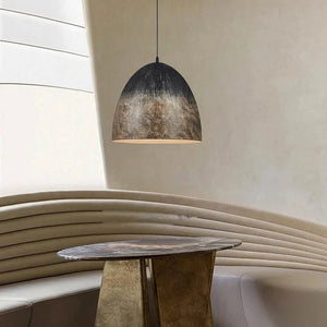 A modern dome-shaped pendant light with a textured, metallic bronze and black finish hanging in a contemporary dining or lounge setting with a curved beige bench and a round table.