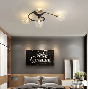 A modern three-light ceiling fixture with black metal arms and clear glass shades is installed in a bedroom with a gray wall, wooden headboard, and framed wall art.