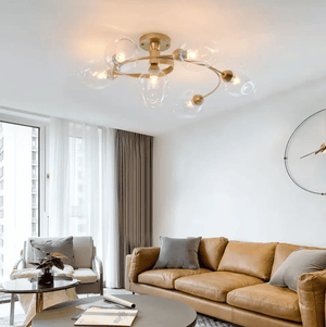 A modern brass ceiling light fixture with six clear glass bubble shades is shown installed in a bright living room with a tan leather sofa, gray pillows, and a large round wall clock.
