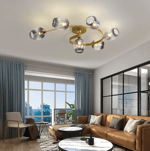 Modern multi-light chandelier with six glass shades and a gold-toned circular frame, installed on a ceiling in a contemporary living room with a brown leather sofa, blue curtains, and large windows.