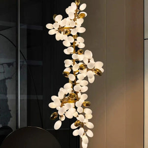 A modern vertical chandelier with a gold branch-like metal frame and numerous oval white frosted glass petals, illuminated against a dark and warm interior background.