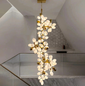 A modern multi-light chandelier with frosted white flower-shaped bulbs and gold metal accents, hanging in a contemporary home interior with a glass railing and geometric ceiling.