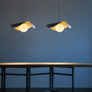 Two modern pendant lights with overlapping leaf-like shades in black, gold, and white, hanging above a minimalist table against a dark blue wall.
