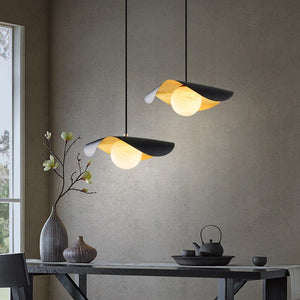 Two modern pendant lights with black and gold leaf-shaped shades hang over a dark table in a minimalist dining room setting with a textured gray wall.