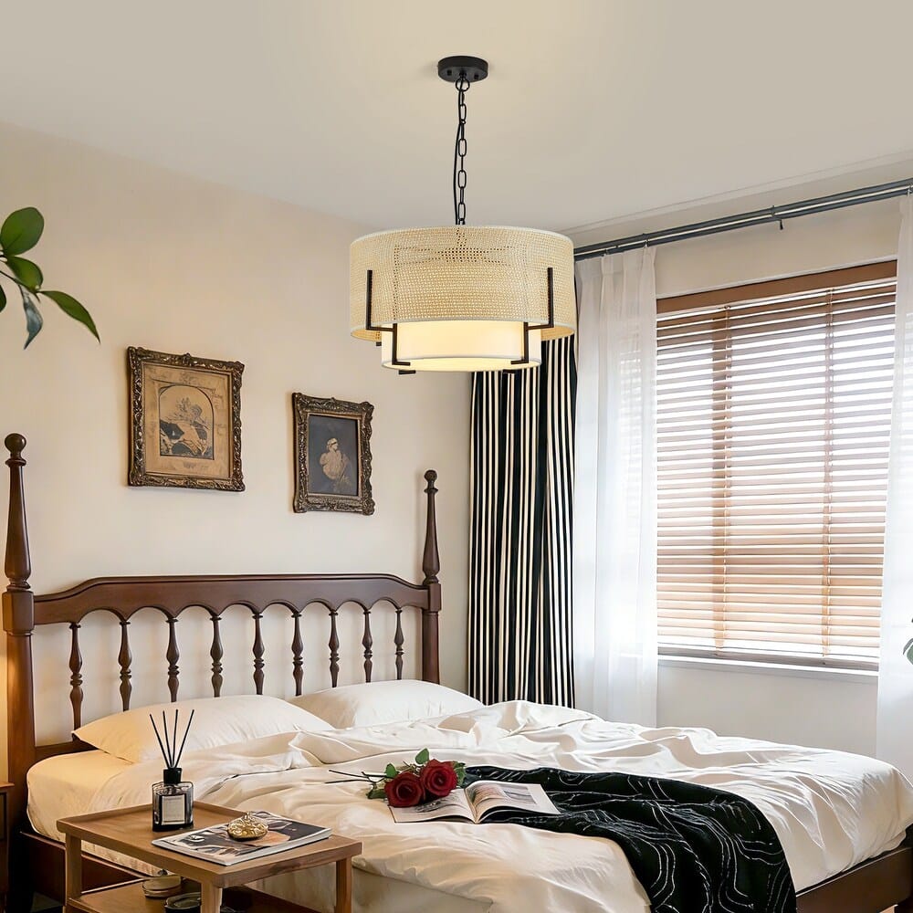 A modern drum pendant light with a cream woven shade and black metal frame hangs over a bed in a styled bedroom with white bedding, wooden headboard, striped curtains, and a window with blinds.