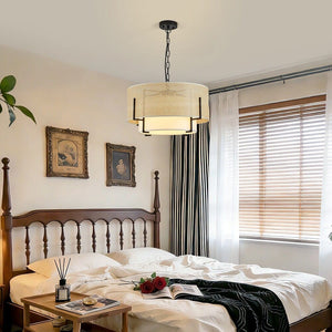 A modern drum pendant light with a cream woven shade and black metal frame hangs over a bed in a styled bedroom with white bedding, wooden headboard, striped curtains, and a window with blinds.