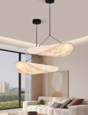 A modern two-light pendant chandelier with organic, flowing shell-shaped shades made of a textured beige material, suspended by black cords from a black ceiling canopy in a bright, contemporary living room.