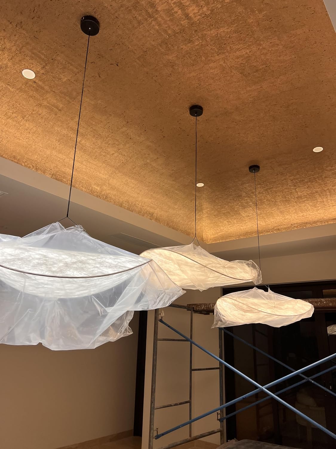 Three modern pendant light fixtures wrapped in protective plastic film, hanging from a textured gold ceiling in an interior room with scaffolding visible below.