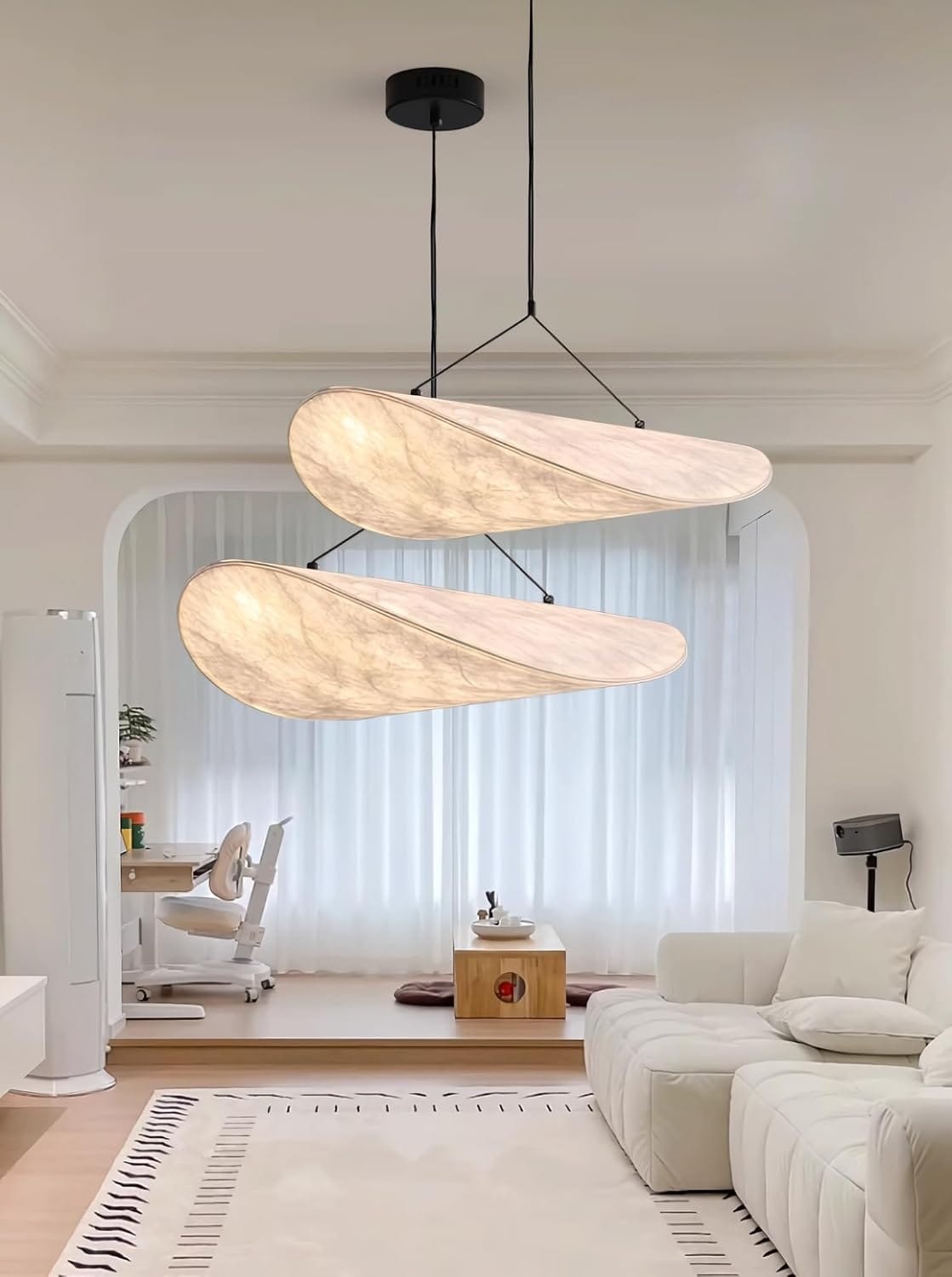 A modern two-light pendant chandelier with organic, wing-like shapes made of warm cream-colored stone or marble, hanging in a bright, contemporary living room with a white sofa and large windows.