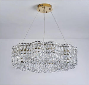 A modern oval chandelier with a brushed gold ceiling canopy and suspension rods, featuring a clear faceted crystal glass body that reflects light. The chandelier is shown hanging against a plain white ceiling and wall.