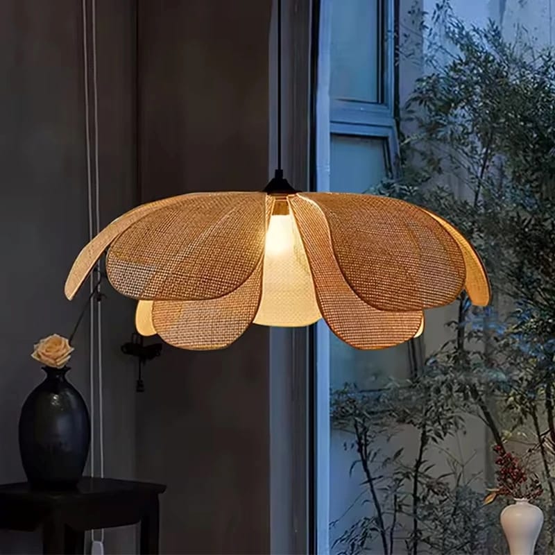 A flower-shaped pendant lamp with woven natural-fiber petals and a glowing warm bulb in the center, hanging in a dimly lit interior near a window.