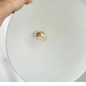 A close-up of a hand holding the edge of a large, round, off-white ceiling light fixture, showing a clear glass globe bulb with a warm filament at the center.