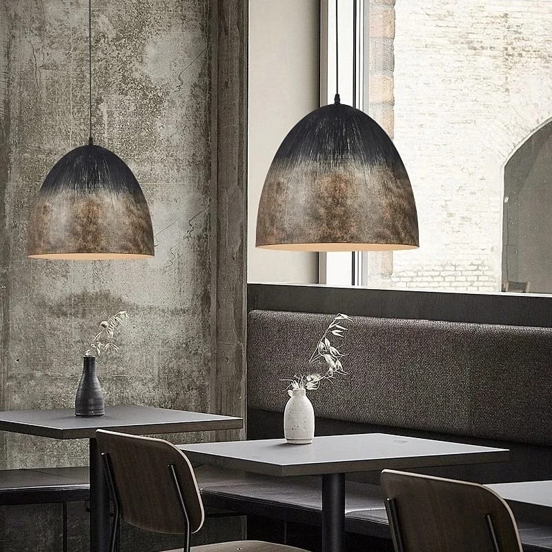 Two dome-shaped pendant lights with a distressed black and bronze metallic finish hang over modern dining tables in a rustic, industrial-style restaurant setting.