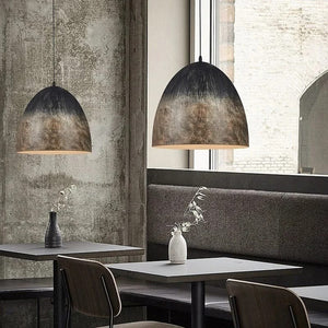 Two dome-shaped pendant lights with a distressed black and bronze metallic finish hang over modern dining tables in a rustic, industrial-style restaurant setting.