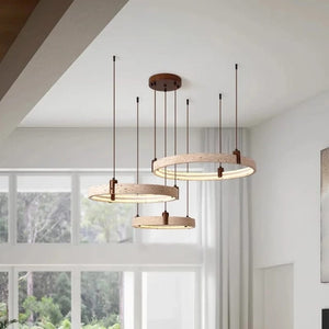 Three modern circular pendant lights with light wood rings and dark metal suspension rods hanging from a white ceiling in a bright room with large windows.