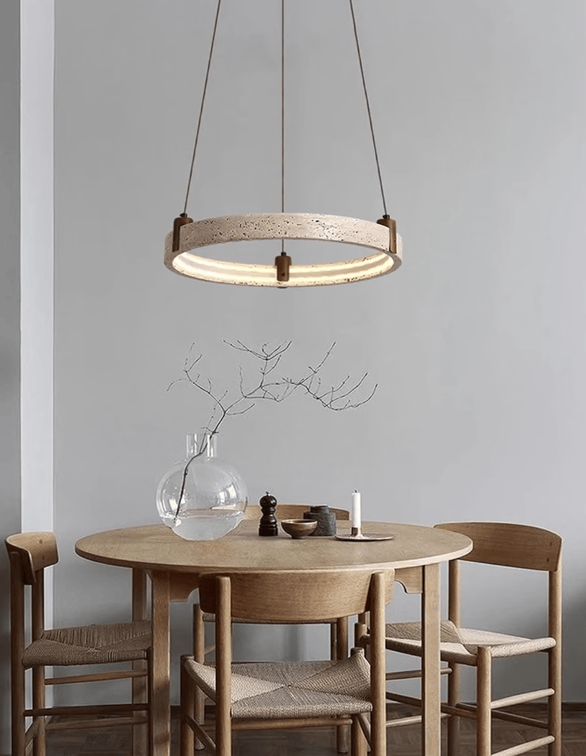 A modern circular pendant light with a light wood ring and warm glowing LED bulbs, hanging above a round wooden dining table with woven chair seats in a minimalist dining room.