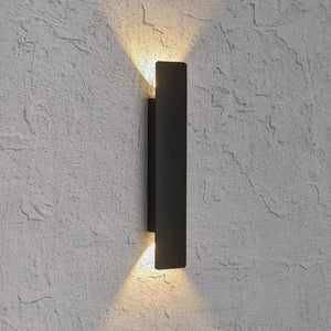 A modern matte black rectangular wall sconce mounted on a textured white stucco wall, casting a warm yellow light upward and downward.