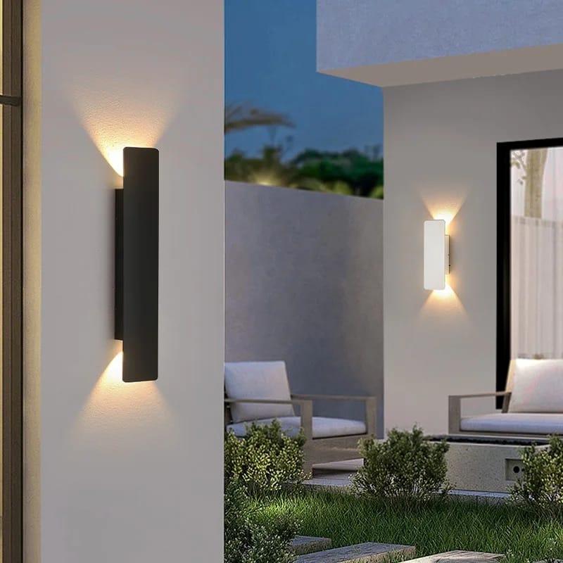 Modern outdoor wall lights on a white wall with a patio and chairs in the background.