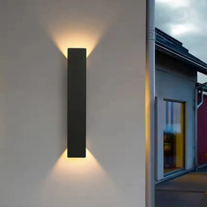 A modern rectangular black wall light fixture mounted on a beige exterior wall, casting a warm yellow light upward and downward. To the right, a narrow view of a house facade and cloudy sky is visible.