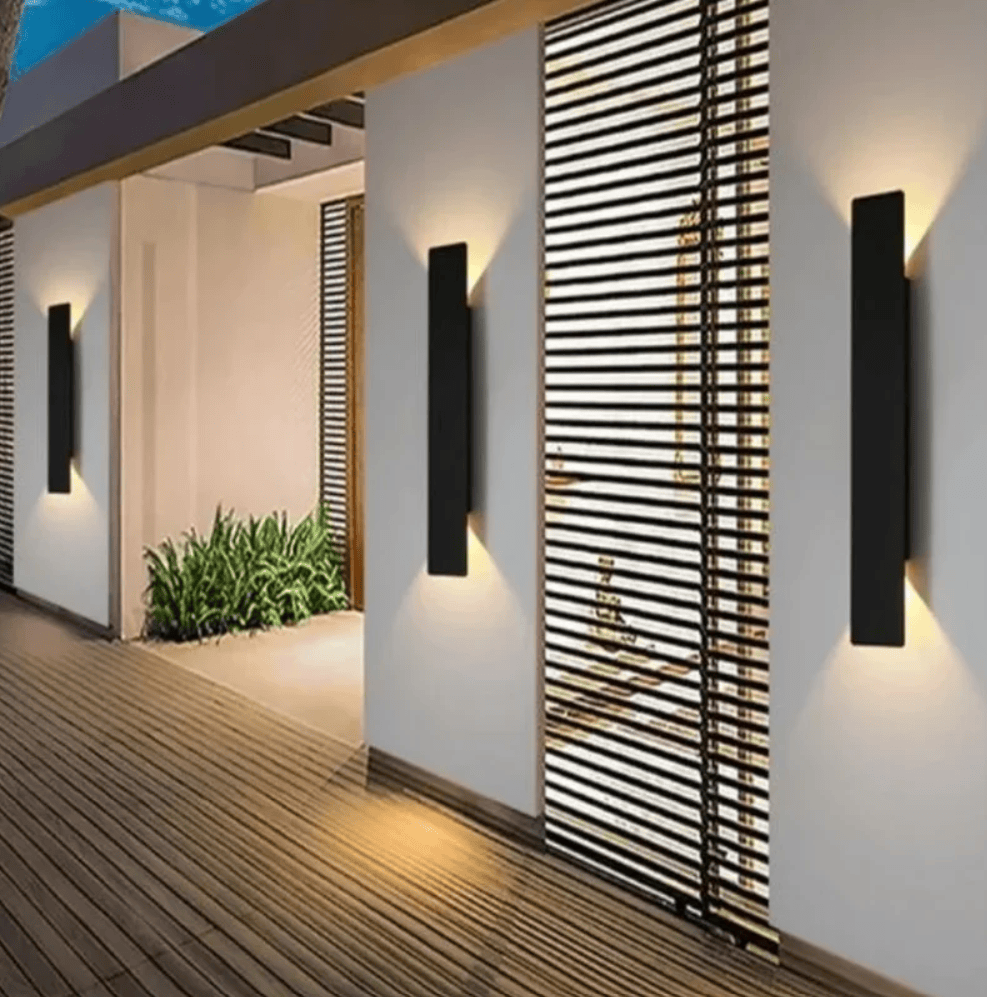 Modern black rectangular wall sconces mounted on white exterior walls, casting warm light onto a wooden deck and slatted screen in an outdoor patio setting.