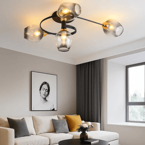 Modern multi-light ceiling fixture with four clear glass shades and a black spiral frame, mounted on a white ceiling in a contemporary living room with a beige sofa and gray curtains.