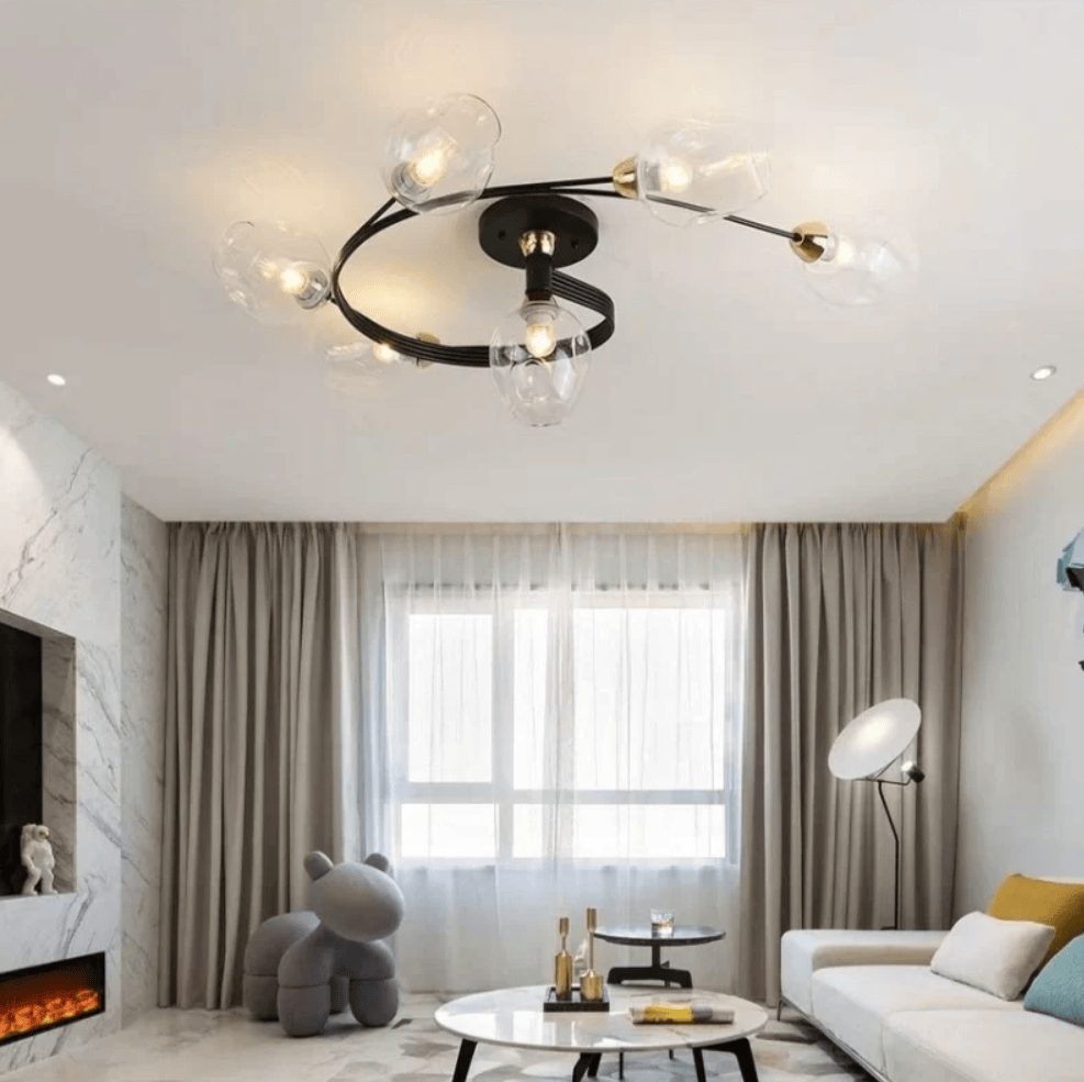 Modern ceiling light fixture with a black circular frame and six clear glass globe bulbs, installed in a contemporary living room with a white sofa, beige curtains, and a fireplace.