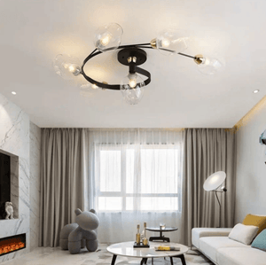 Modern ceiling light fixture with a black circular frame and six clear glass globe bulbs, installed in a contemporary living room with a white sofa, beige curtains, and a fireplace.