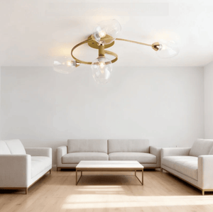 A modern ceiling light fixture with a gold-toned circular frame and three clear glass shades, installed in a bright, minimalist living room with white sofas and light wood flooring.