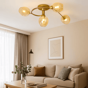 A modern ceiling light fixture with a gold brass frame and four warm, glowing frosted glass globes, installed in a bright, contemporary living room with a beige sofa and neutral-toned walls.