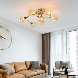 A modern ceiling light fixture with six clear glass globes on a gold-toned metal frame, installed in a bright living room above a tan leather sofa.