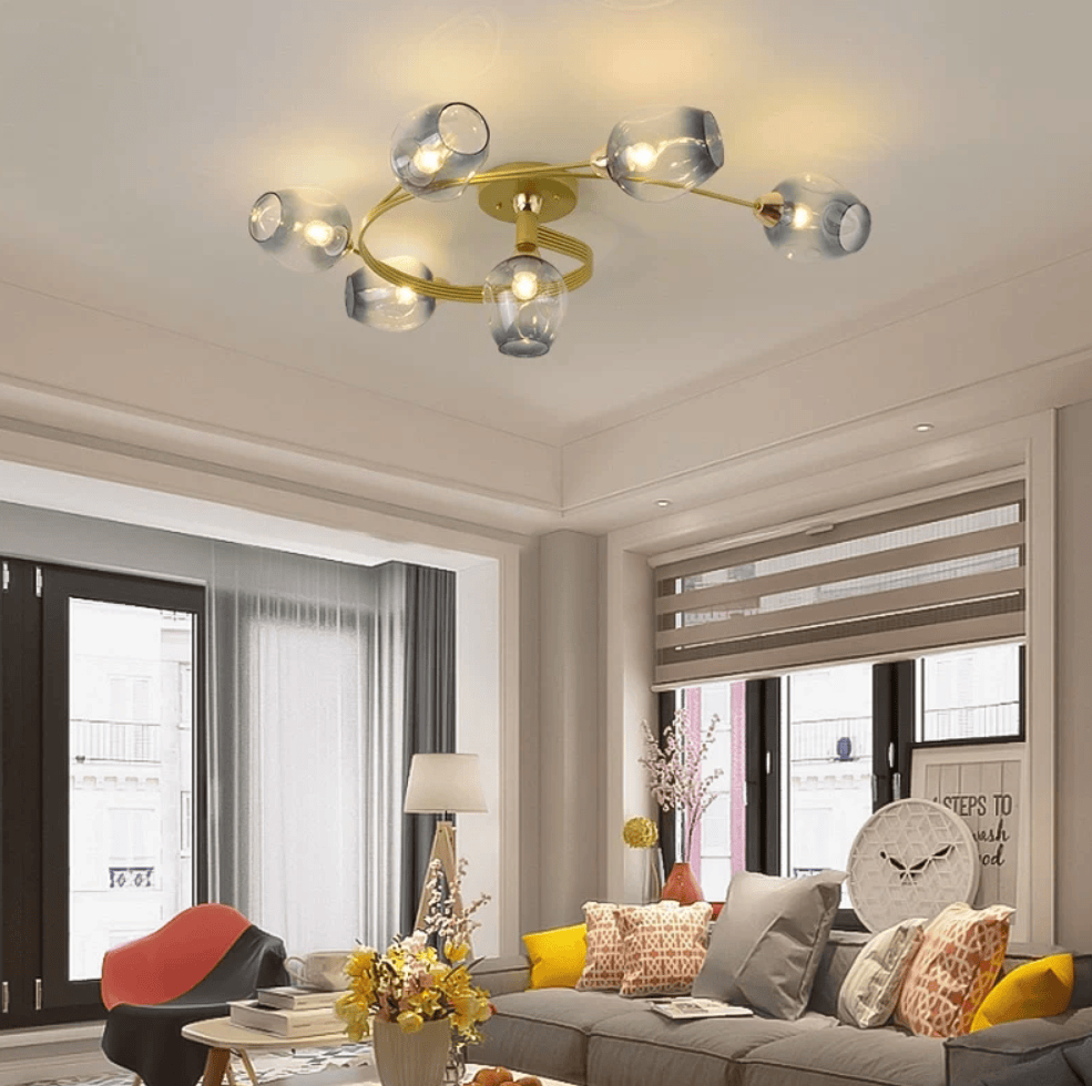 A modern ceiling light fixture with six clear glass shades and a gold-toned circular frame, mounted on the ceiling in a contemporary living room with a gray sofa, yellow accent pillows, and large windows.