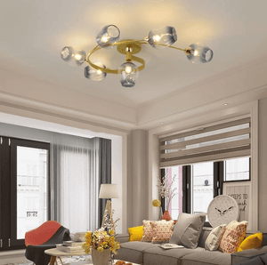 A modern ceiling light fixture with six clear glass shades and a gold-toned circular frame, mounted on the ceiling in a contemporary living room with a gray sofa, yellow accent pillows, and large windows.
