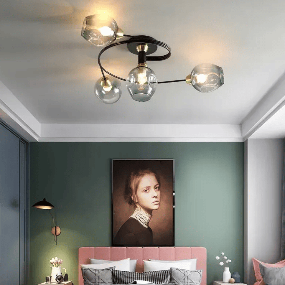 Modern ceiling light fixture with four clear glass globes and a black curved frame, installed in a bedroom with a green wall and pink bed.