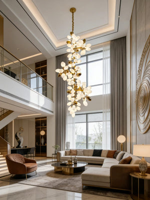 A modern multi-light chandelier with gold-toned metal and numerous frosted white glass flower-like bulbs hangs in a high-ceilinged contemporary living room with a sectional sofa, large windows, and a staircase.