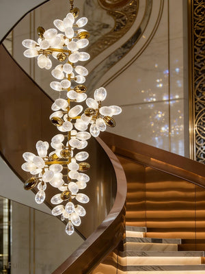 A luxury chandelier with a gold-tone metal frame and clear glass flower-like bulbs, hanging in front of a modern curved staircase with warm lighting.