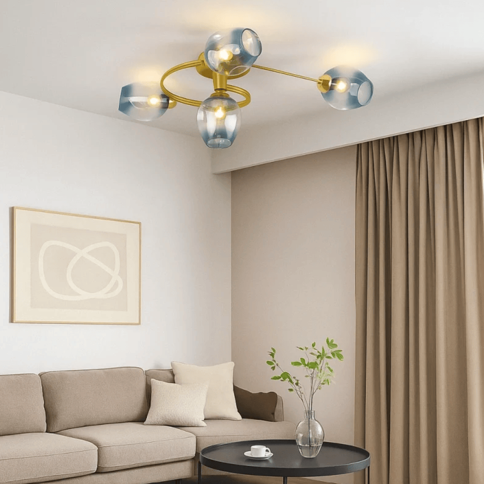 Modern ceiling light fixture with a gold metal frame and four translucent blue glass shades, shown installed in a contemporary living room with a beige sofa, curtains, and a small round table below.