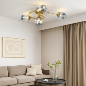 Modern ceiling light fixture with a gold metal frame and four translucent blue glass shades, shown installed in a contemporary living room with a beige sofa, curtains, and a small round table below.