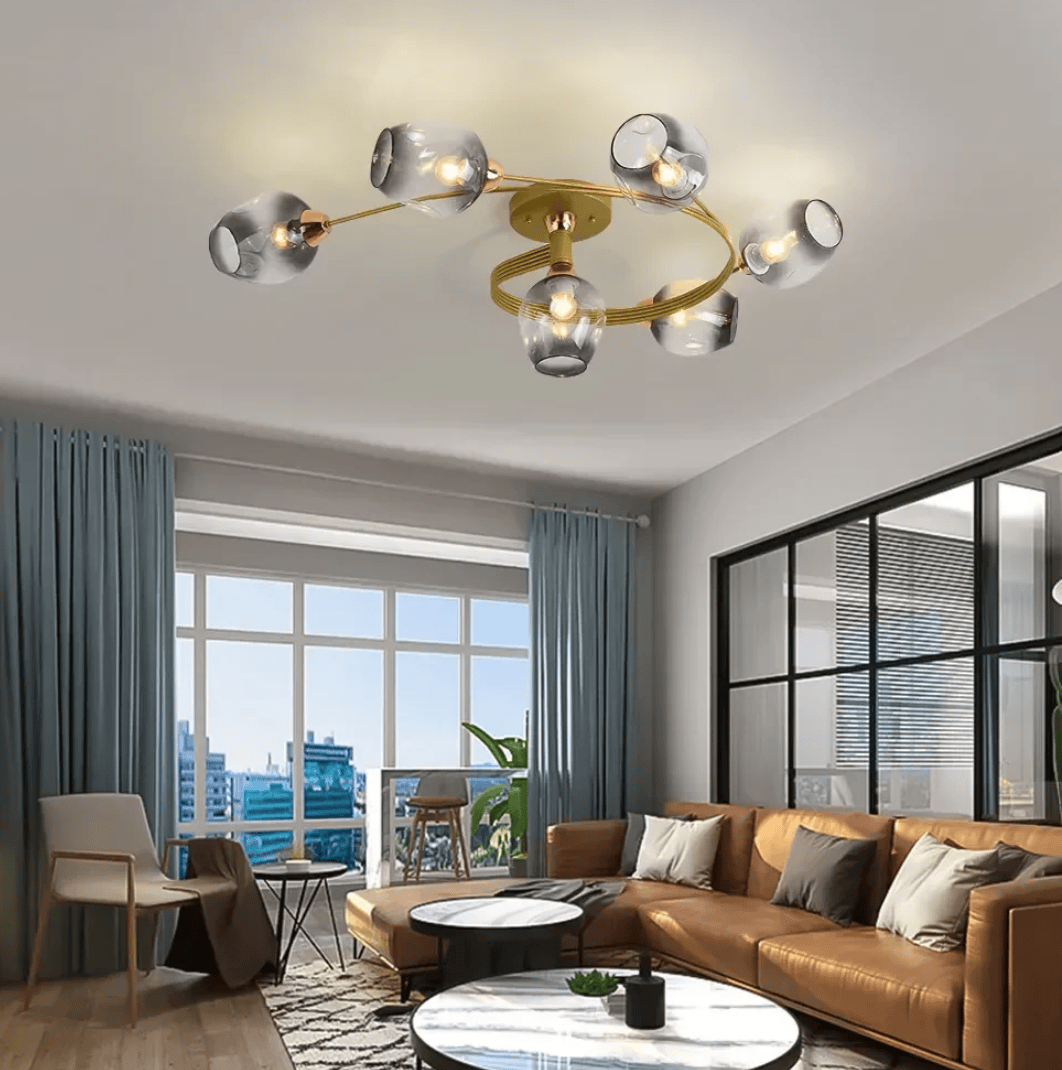 Modern multi-light chandelier with six glass shades and a gold-toned circular frame, installed on a ceiling in a contemporary living room with a brown leather sofa, blue curtains, and large windows.