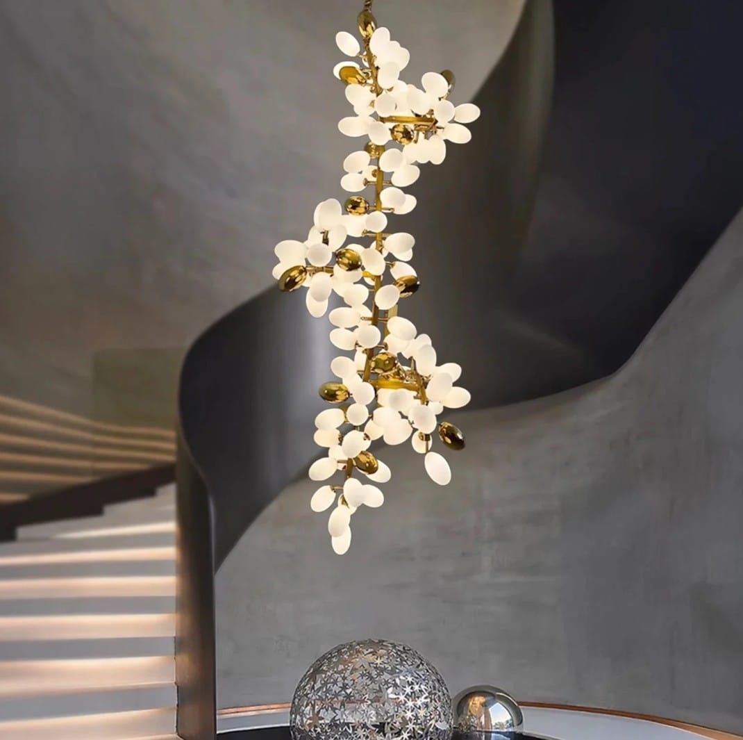 A modern hanging pendant light fixture with white petal-shaped shades and a gold branch-like frame, shown suspended in a contemporary interior with curved gray walls and a staircase in the background.