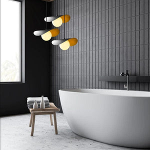 Modern freestanding bathtub in a contemporary bathroom with dark gray wall tiles, a small wooden stool with folded towels, and a multi-light pendant fixture overhead.
