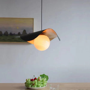 A modern wavy pendant light with a black and brushed gold metal shade and a glowing warm white globe bulb, hanging over a table with a plate of grapes in a minimalist room setting.