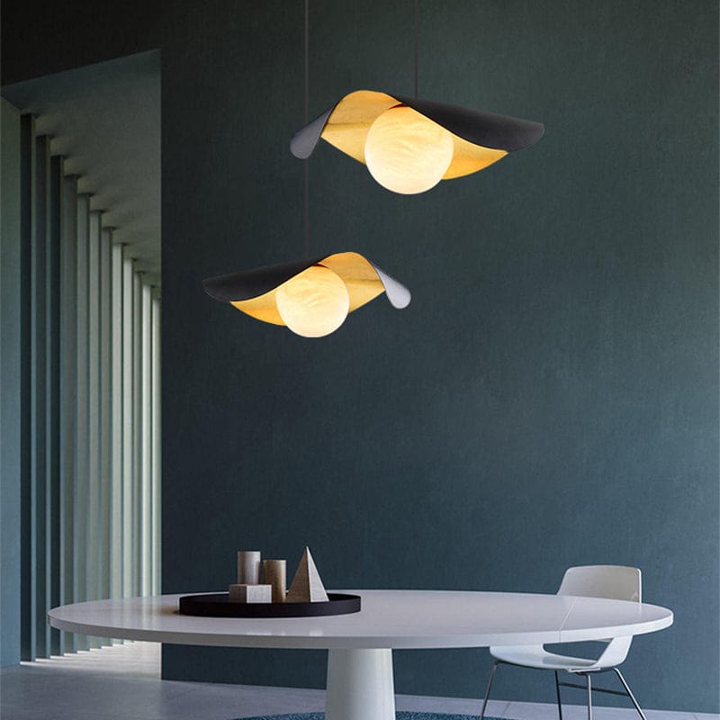Two modern pendant lights with curved black and warm yellow-gold shades hang above a white dining table in a minimalist room with a dark teal wall.