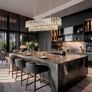 A modern rectangular crystal chandelier with a warm light glowing from within, hanging in a contemporary kitchen with a marble island and dark cabinetry.