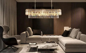 A modern living room featuring a large rectangular crystal chandelier hanging above a light gray sectional sofa and marble coffee table, with a dark wood slatted accent wall behind.