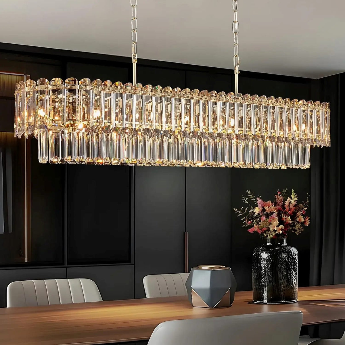 A rectangular crystal chandelier with multiple tiers of hanging clear glass rods and warm illuminated bulbs, suspended over a dining table in a modern dark interior.