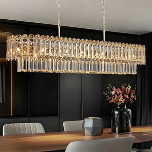 A rectangular crystal chandelier with multiple tiers of hanging clear glass rods and warm illuminated bulbs, suspended over a dining table in a modern dark interior.