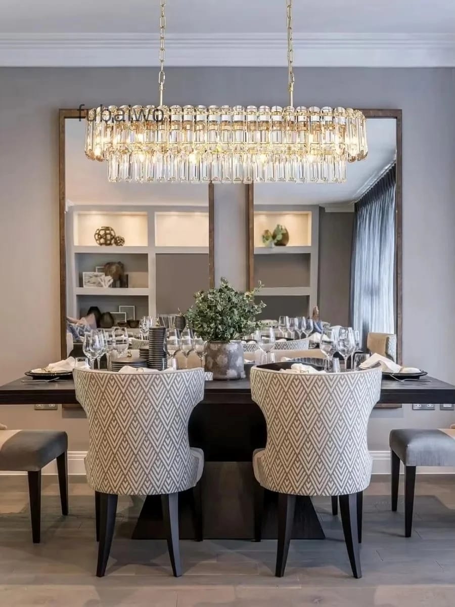 A modern dining room featuring a large rectangular gold-framed chandelier with hanging clear crystal prisms, suspended over a dark wood dining table set with patterned beige chairs.